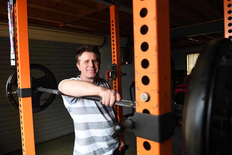 Image of a man leaning on a bench press with large weights and smiling at the camera