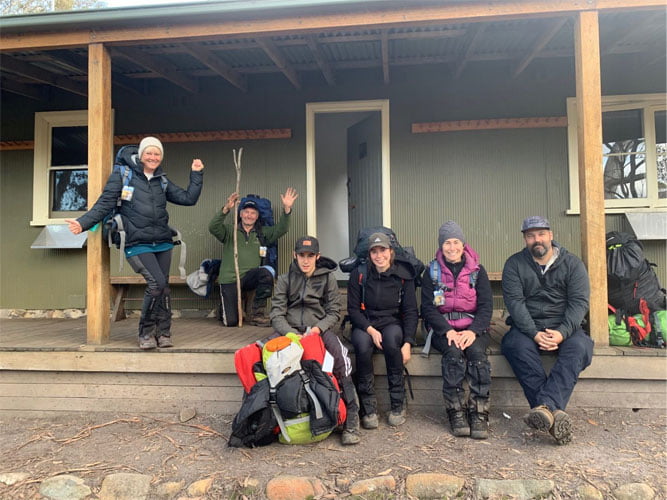 Group of hikers facing the camera and smiling, while sitting on the veranda of a hiking hut with backpacks.